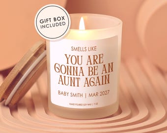 May include: A frosted glass candle with a lit wick and a bamboo lid. The label reads: SMELLS LIKE YOU ARE GONNA BE AN AUNT AGAIN, BABY SMITH | MAR 2027, HAND POURED SOY WAX | 7 OZ. A circular sticker on the glass states: GIFT BOX INCLUDED.