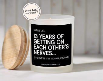 May include: A frosted glass candle with a black label featuring the white text: SMELLS LIKE 13 YEARS OF GETTING ON EACH OTHER'S NERVES... (AND WE'RE STILL GOING STRONG) HAND POURED SOY WAX | 7 OZ. A round wooden lid sits beside the candle, and a circular tag reads GIFT BOX INCLUDED.