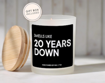 May include: A frosted glass candle with a black label featuring the white text SMELLS LIKE 20 YEARS DOWN and HAND POURED SOY WAX | 7 OZ. A round bamboo lid sits to the left. A circular badge at the top left reads GIFT BOX INCLUDED in black text.