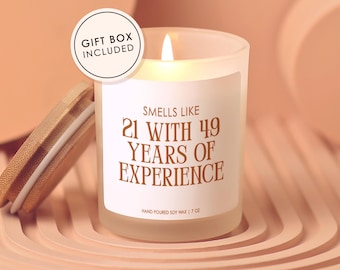 May include: A lit, frosted glass candle with a wooden lid, labeled "Smells Like 21 With 49 Years of Experience." The candle is 7 oz and comes in a gift box. The background is a light peach color.