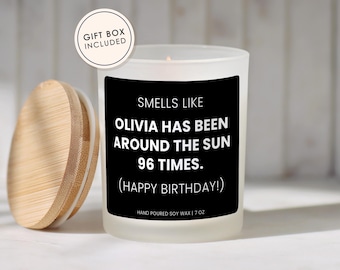 May include: A frosted glass candle with a black label that reads: SMELLS LIKE OLIVIA HAS BEEN AROUND THE SUN 96 TIMES. (HAPPY BIRTHDAY!) HAND POURED SOY WAX | 7 OZ. A round bamboo lid sits to the left. A circular badge states GIFT BOX INCLUDED.