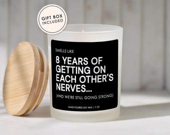 May include: A frosted glass candle with a black label that reads: SMELLS LIKE 8 YEARS OF GETTING ON EACH OTHER'S NERVES... (AND WE'RE STILL GOING STRONG) HAND POURED SOY WAX | 7 OZ. A round bamboo lid sits to the left, and a circular tag says GIFT BOX INCLUDED.