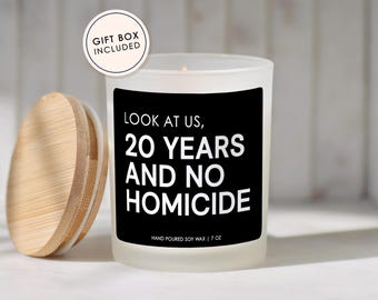 May include: A frosted glass candle with a black label featuring the white text LOOK AT US, 20 YEARS AND NO HOMICIDE and HAND POURED SOY WAX | 7 OZ. A circular bamboo lid sits to the left, and a round sticker reads GIFT BOX INCLUDED at the top left.
