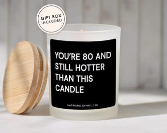 May include: A white candle with a black label that reads "YOU'RE 80 AND STILL HOTTER THAN THIS CANDLE." The candle has a wooden lid and is labeled "HAND POURED SOY WAX | 7 OZ." A gift box is included.