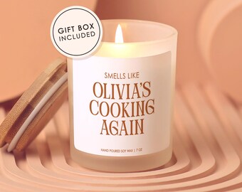 May include: A frosted glass candle with a lit wick and a bamboo lid. The white label features the brown text 'SMELLS LIKE OLIVIA'S COOKING AGAIN' and 'HAND POURED SOY WAX | 7 OZ'. A circular badge on the top left reads 'GIFT BOX INCLUDED' in black text.