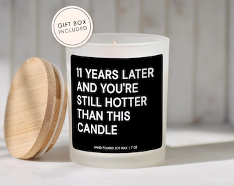 May include: A frosted glass candle with a black label featuring the white text 11 YEARS LATER AND YOU'RE STILL HOTTER THAN THIS CANDLE and HAND POURED SOY WAX | 7 OZ. A round wooden lid sits to the left, and a circular badge reads GIFT BOX INCLUDED.