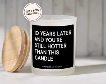May include: A white frosted glass candle with a black label that reads "10 YEARS LATER AND YOU'RE STILL HOTTER THAN THIS CANDLE." The candle has a wooden lid and is labeled "HAND POURED SOY WAX | 7 OZ." A gift box is included.