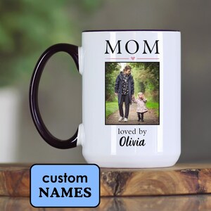 May include: White ceramic mug with a black handle and rim. The mug features a photo of a person and child, with the word "MOM" above the photo and "loved by Olivia" below. A blue rectangle at the bottom says "custom NAMES".