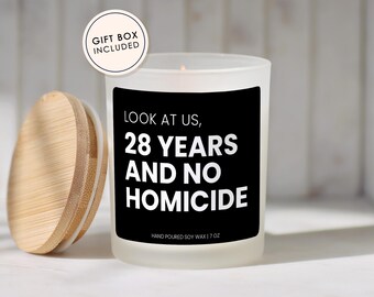 May include: A frosted glass candle with a black label featuring the white text LOOK AT US, 28 YEARS AND NO HOMICIDE and HAND POURED SOY WAX | 7 OZ. The candle has a light wood lid and a circular sticker that reads GIFT BOX INCLUDED.