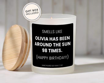 May include: A frosted glass candle with a black label that reads SMELLS LIKE OLIVIA HAS BEEN AROUND THE SUN 98 TIMES. (HAPPY BIRTHDAY!) HAND POURED SOY WAX | 7 OZ. A round wooden lid sits to the left. A circular badge states GIFT BOX INCLUDED.