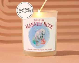 May include: A lit candle in a frosted glass jar with a label that reads "Smells Like Manatee Hugs." The label features a watercolor illustration of a manatee. The text "Gift Box Included" is in a circle above the candle.