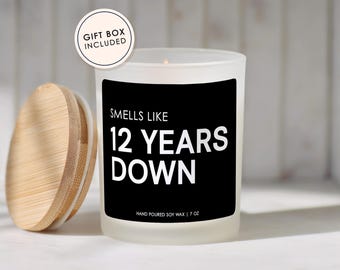 May include: A frosted glass candle with a black label that reads "SMELLS LIKE 12 YEARS DOWN" and "HAND POURED SOY WAX | 7 OZ". A round wooden lid sits to the left of the candle. A circular badge in the upper left corner states "GIFT BOX INCLUDED".