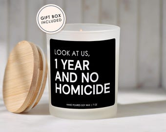 May include: A white frosted glass candle with a black label that reads "LOOK AT US, 1 YEAR AND NO HOMICIDE." The candle has a wooden lid and is labeled "HAND POURED SOY WAX | 7 OZ." A circular sticker reads "GIFT BOX INCLUDED."