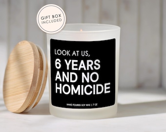May include: A frosted glass candle with a black label featuring the white text LOOK AT US, 6 YEARS AND NO HOMICIDE and HAND POURED SOY WAX | 7 OZ. A round bamboo lid sits to the left, and a circular badge reads GIFT BOX INCLUDED at the top left.