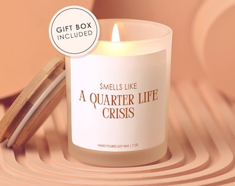 May include: A lit soy wax candle in a frosted glass jar with a wooden lid. The label reads "Smells Like A Quarter Life Crisis." The candle is 7 oz and comes in a gift box.