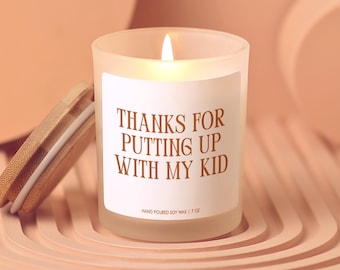 May include: A lit soy wax candle in a frosted glass jar with a wooden lid. The label reads "THANKS FOR PUTTING UP WITH MY KID." The candle is 7 oz.