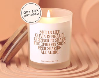 May include: A lit, frosted glass candle with a wooden lid. The candle has a white label with the text "SMELLS LIKE OLIVIA IS FINALLY LICENSED TO SHARE THE OPINIONS SHE'S BEEN SHARING ALL ALONG." The candle is 7 oz and includes a gift box.