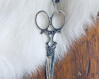 Op de afbeelding: Een zilverkleurige schaar hanger ketting met een gedetailleerd ontwerp. De hanger hangt aan een zwarte ketting.