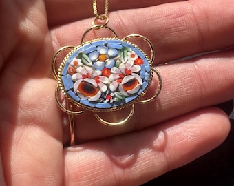 May include: A gold-tone pendant necklace featuring an oval micro-mosaic centerpiece. The mosaic displays a floral pattern with light blue, white, red, and green glass tiles. The pendant is framed by a gold-tone scalloped wire border and hangs from a thin gold-tone chain.