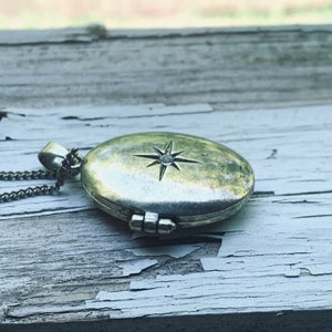 May include: A silver locket necklace with a star design on the front. The locket is open and the inside is visible. The necklace is on a wooden surface.
