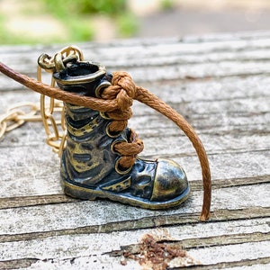 Può includere: Una collana con catena dorata con un ciondolo a forma di stivale dettagliato. Lo stivale è in metallo e ha un laccio in pelle marrone legato intorno.