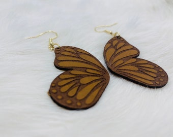 Puede incluir: Un par de pendientes de mariposa marrones con ganchos dorados. Los pendientes están hechos de cuero y tienen un diseño detallado.