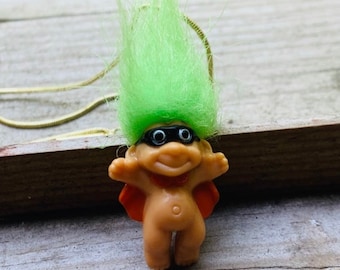 May include: A green haired troll wearing black eye mask and a red cape, is a pendant on a gold chain.