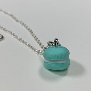Puede incluir: Un collar de cadena plateada con un dije de macaron azul claro. El macaron tiene un relleno blanco.
