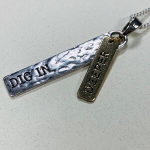 Op de afbeelding: Zilveren ketting met twee hangers. De grotere hanger is zilver en zegt "DIG IN". De kleinere hanger is goud en zegt "DEEPER".