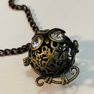 Può includere: Un ciondolo a forma di gufo in bronzo con accenti di cristallo trasparente appeso a una catena. Il ciondolo è decorato con intricati disegni a filigrana.
