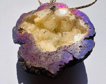 Peut inclure: Un collier pendentif composé d'une tranche de géode brute avec un bord extérieur métallique irisé violet et bleu, entourant un amas de cristaux jaune pâle translucides au centre, suspendu à une fine chaîne en métal doré.