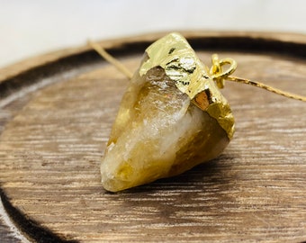 Puede incluir: Un colgante de cristal de cuarzo transparente chapado en oro con una anilla dorada. El cristal tiene una forma natural irregular y un tono amarillo claro.