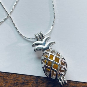 Könnte beinhalten: Eine silberne Kettenkette mit einem ananasförmigen Anhänger. Der Anhänger ist ein Käfig mit einer gelben Mitte.