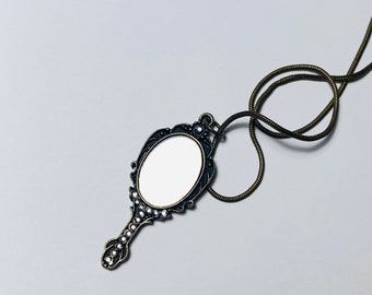 May include: A gold-toned necklace with a decorative mirror pendant. The mirror is oval-shaped and has a black frame with ornate details. The necklace is made of a thin, braided chain.