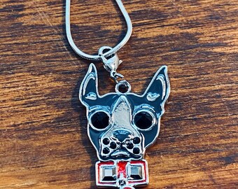 Può includere: Una collana con catena d'argento con un ciondolo a forma di cane nero e argento. Il cane ha gli occhi rossi e un papillon rosso con un gioiello rosso al centro.