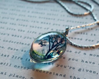 Puede incluir: Un collar de cadena de plata con un colgante redondo de vidrio que presenta una imagen pintada a mano de un árbol con un fondo de cielo azul y hierba verde. El colgante tiene un diámetro de aproximadamente 2,5 cm.