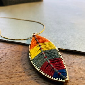 May include: A gold chain necklace with a colorful, woven feather pendant. The feather is made of red, orange, yellow, green, and blue threads.
