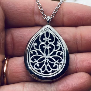 Könnte beinhalten: Eine silberne, tropfenförmige Anhänger-Halskette mit einem filigranen Design. Der Anhänger hat einen schwarzen Hintergrund und ein filigranes Muster in Silber.