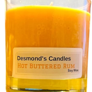 May include: A clear glass candle holder filled with a vibrant orange candle. The candle has a label that reads "Desmond's Candles Hot Buttered Rum Soy Wax". The candle is lit and has a visible wick.