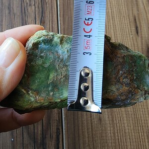Może przedstawiać: Szorstki, zielony kamień jest mierzony za pomocą miarki. Kamień ma około 3,8 cm wysokości. Kamień ma nakrapiany wygląd z odcieniami zieleni i brązu.