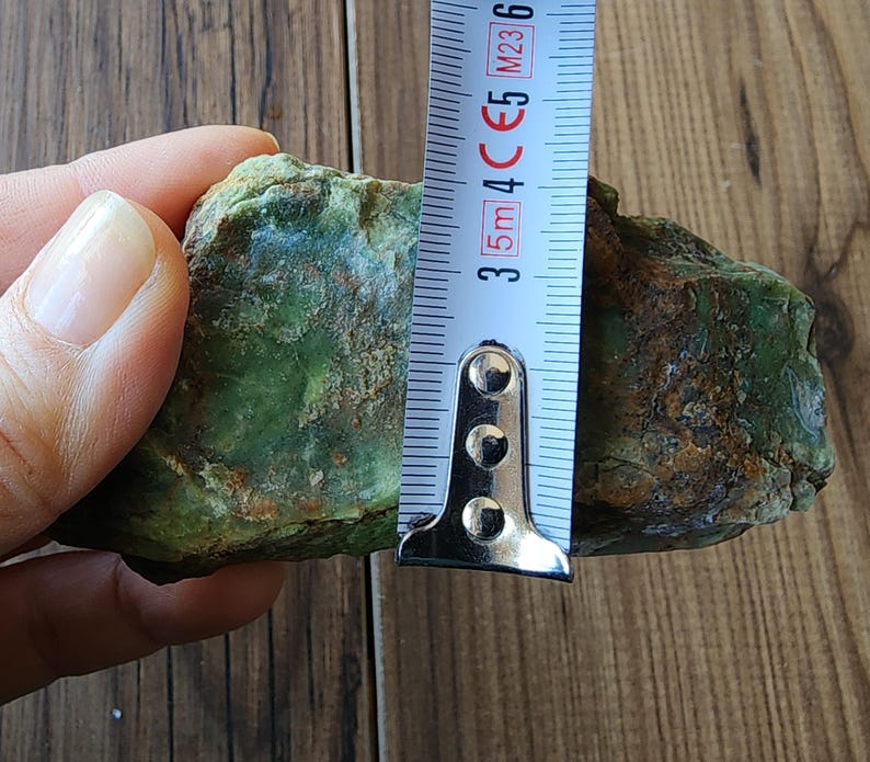 Może przedstawiać: Szorstki, zielony kamień jest mierzony za pomocą miarki. Kamień ma około 3,8 cm wysokości. Kamień ma nakrapiany wygląd z odcieniami zieleni i brązu.
