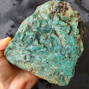 May include: A rough, irregularly shaped rock with a vibrant teal and green color palette. The stone's surface is textured with visible cracks and layers, showcasing a natural, raw aesthetic. The rock is held in a hand, highlighting its size and form.