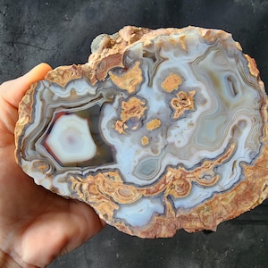 May include: A polished agate stone with intricate patterns of white, gray, brown, and orange hues. The stone's center features a white and red circular design, surrounded by layered bands. The rough edges of the stone are visible, showcasing its natural texture.