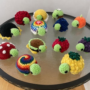 May include: A collection of twelve small, handmade crochet turtles with light green heads and black eyes. Each turtle features a unique shell design, including solid colors, rainbow patterns, polka dots, and textured fruit-inspired shapes.