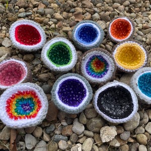 May include: A collection of crocheted geode-shaped objects with various colored interiors, including red, green, blue, orange, yellow, and rainbow patterns. Each geode has a gray and white border, and they are arranged on a bed of small rocks.