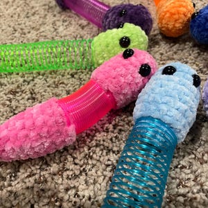 May include: A set of colorful crocheted worm toys with springy bodies. The worms are pink, blue, green, purple, and orange. Each worm has two black eyes.