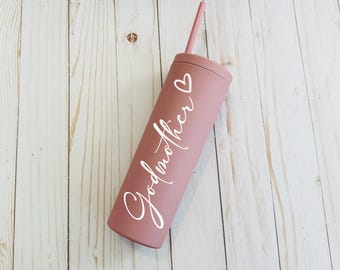May include: A matte dusty rose cylindrical tumbler with a matching lid and straw. The word Godmother is printed in white cursive script vertically along the side, accompanied by a small white heart outline positioned above the text.