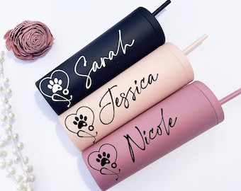 May include: Three cylindrical tumblers with straws in black, light pink, and mauve. Each features a custom name in black script: Sarah, Jessica, and Nicole. Beside each name is a graphic of a heart containing a paw print and a stethoscope.