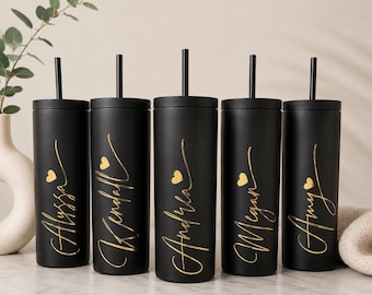May include: Five matte black cylindrical tumblers with lids and matching black straws. Each tumbler features a gold script name and a small gold heart icon. The names displayed are Alyssa, Kendall, Andrea, Megan, and Amy, arranged in a row.
