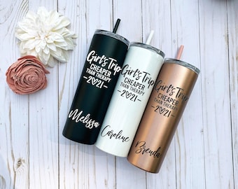 May include: Three tall cylindrical tumblers with lids and straws in black, white, and rose gold. Each features the text Girl's Trip CHEAPER THAN THERAPY 2021 and a unique name: Melissa, Caroline, or Brenda, printed in a black or white script font.